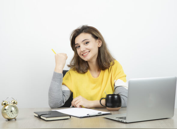 Photo of female office worker sitting at the desk with laptop and clipboard